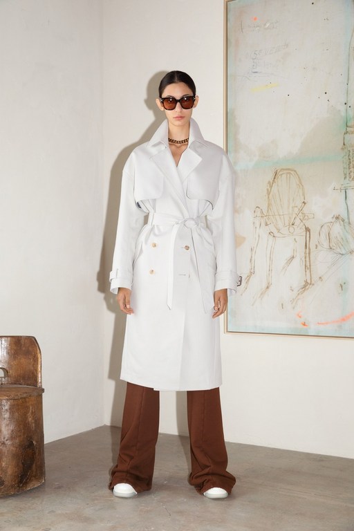Model in white trench coat and brown trousers wearing sunglasses standing against a wall with abstract painting