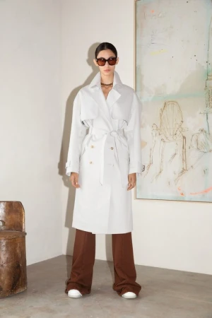 Model in white trench coat and brown trousers wearing sunglasses standing against a wall with abstract painting