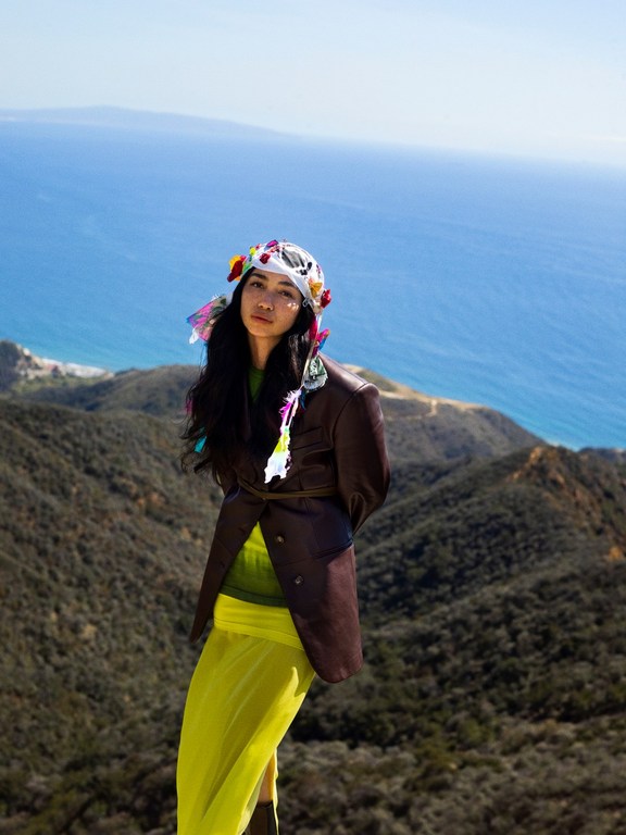 Adult woman in a brown jacket and yellow skirt standing on a hill overlooking the ocean and mountains, wearing a floral headpiece.