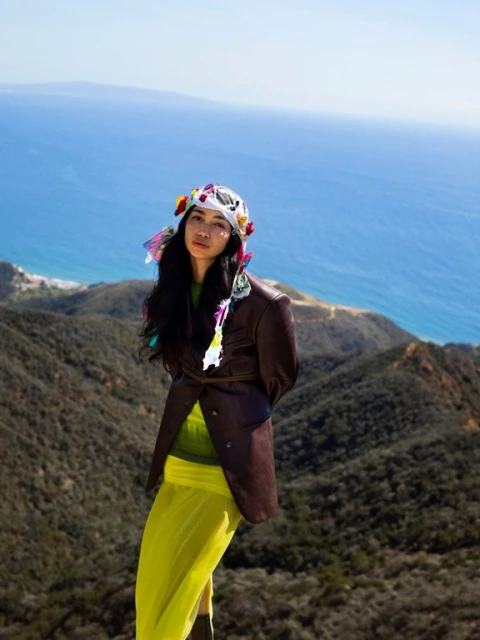 Adult woman in a brown jacket and yellow skirt standing on a hill overlooking the ocean and mountains, wearing a floral headpiece.