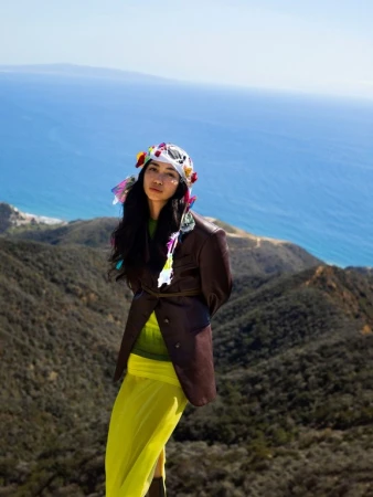 Adult woman in a brown jacket and yellow skirt standing on a hill overlooking the ocean and mountains, wearing a floral headpiece.