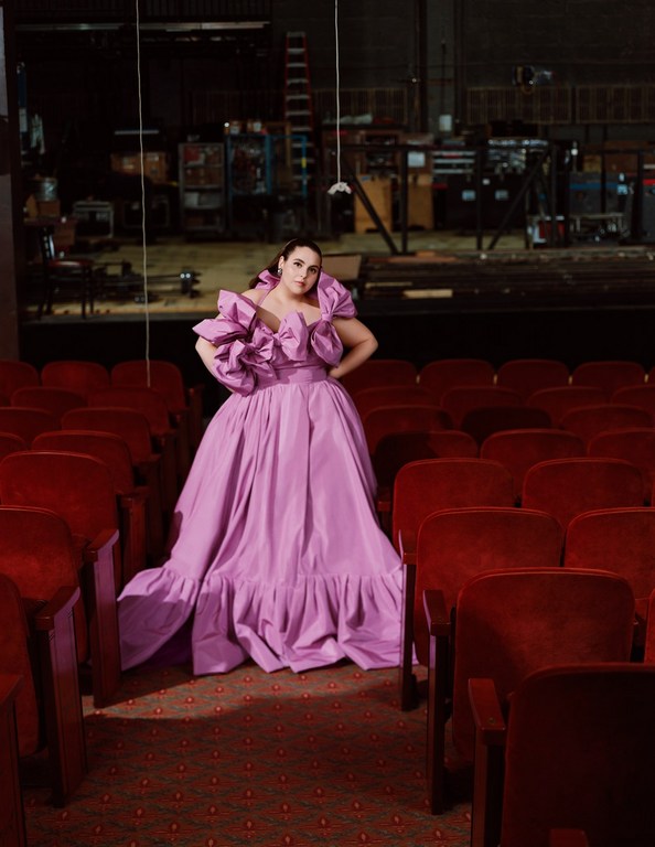 Woman poses in a bright purple gown in an empty theater with red velvet chairs. A stage is visible in the background.