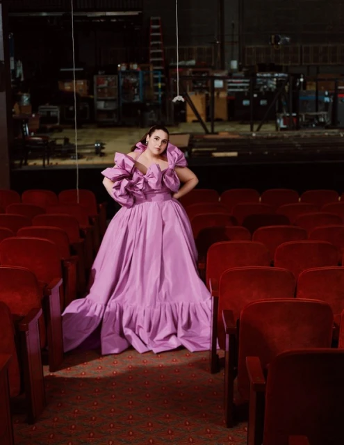 Woman poses in a bright purple gown in an empty theater with red velvet chairs. A stage is visible in the background.