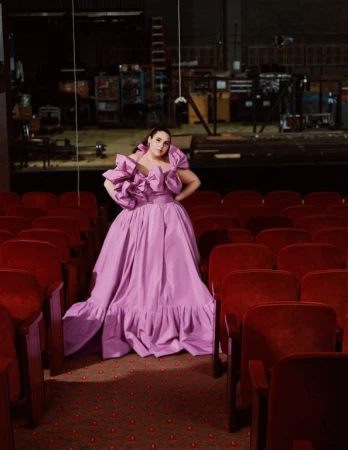 Woman poses in a bright purple gown in an empty theater with red velvet chairs. A stage is visible in the background.