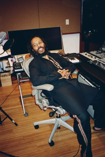Portrait of adult male musician with dreadlocks in black shirt sitting in chair in recording studio in front of mixing console and equipment.
