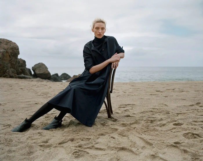 Model wearing dark dress seated on a wooden chair on a sandy beach under an overcast sky.