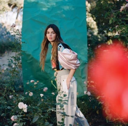 Kendall Jenner standing in a garden with pale pink roses, wearing a color-blocked top and grey belted trousers, styled by Max Ortega.