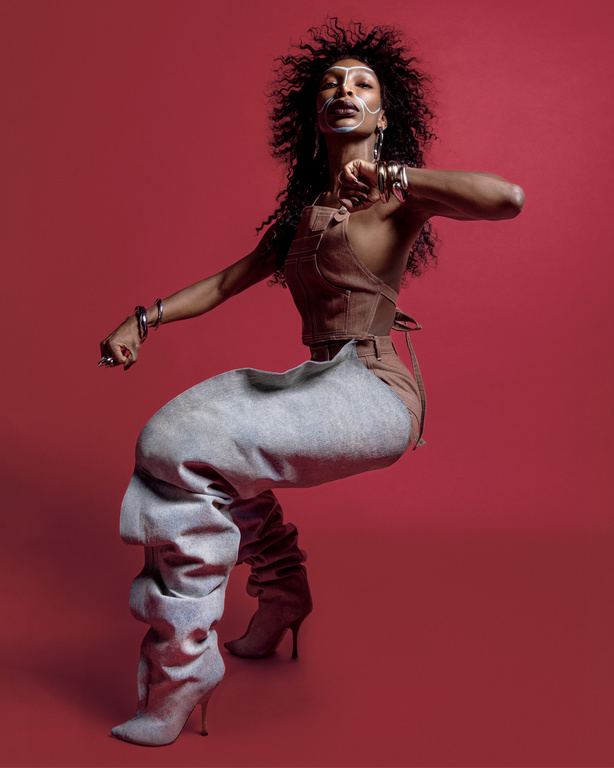 Fashion portrait of a woman in sculptural clothing, posing in a dynamic stance against a red background, with curly hair and face paint.