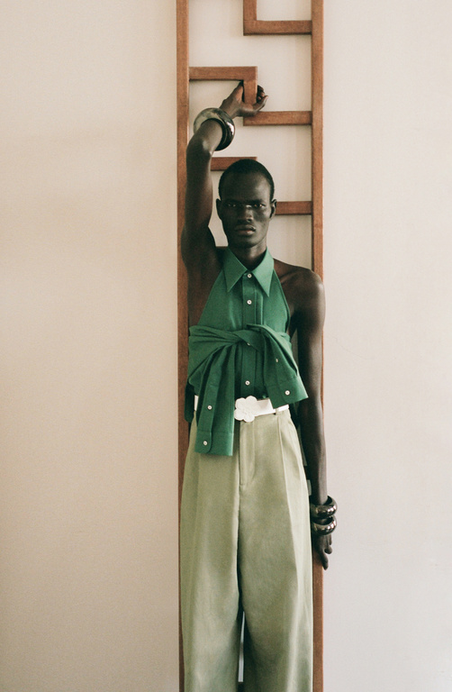 An adult male with dark skin stands in a green shirt and beige pants against a wall with a geometric wooden sculpture.