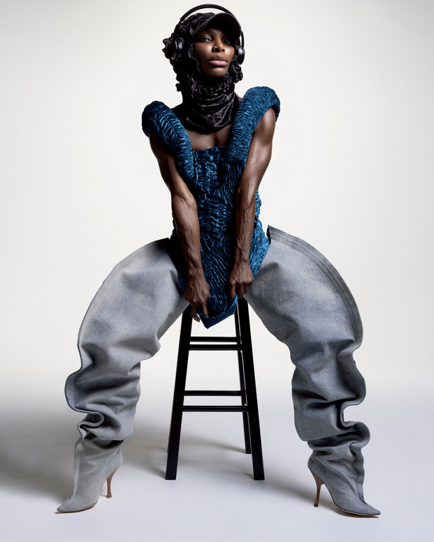 Fashionable Black adult woman wearing an avant-garde outfit with blue top, baggy pants, headphones and cap, sitting on a stool indoors.