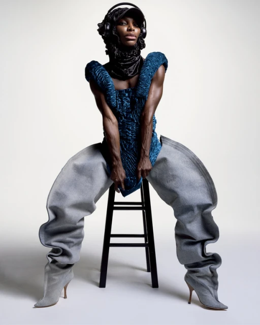 Fashionable Black adult woman wearing an avant-garde outfit with blue top, baggy pants, headphones and cap, sitting on a stool indoors.