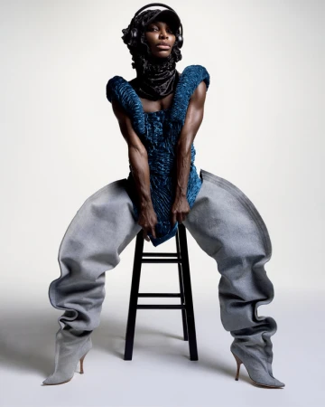 Fashionable Black adult woman wearing an avant-garde outfit with blue top, baggy pants, headphones and cap, sitting on a stool indoors.