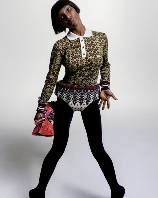 Woman in patterned top and black leggings poses in studio. She holds a red purse and looks at the camera with a slight head tilt.