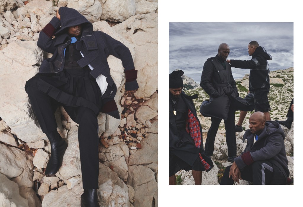 Fashion shoot of a Black model wearing dark clothing posing on rocks, with additional models in background on mountain top.