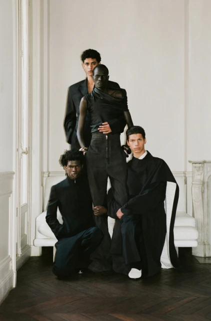 Group portrait of four young adult male fashion models in dark clothing posing indoors against a white wall and minimalist couch.
