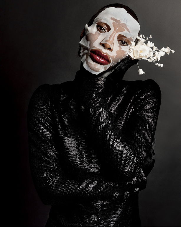 Person in black sequined garment and facial mask poses with white flowers against dark background in beauty editorial photography.