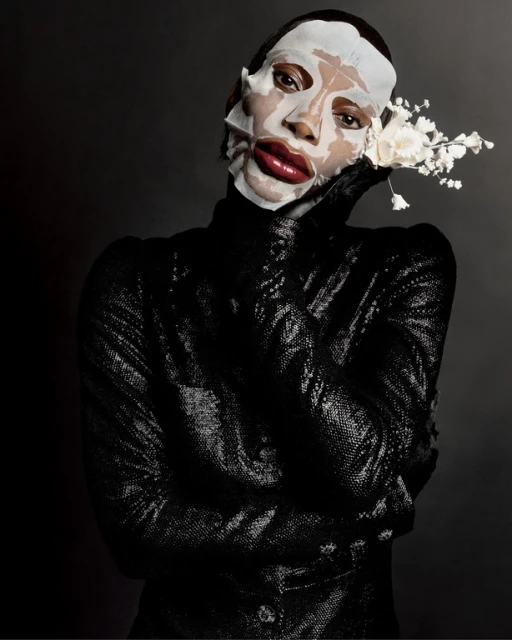 Person in black sequined garment and facial mask poses with white flowers against dark background in beauty editorial photography.