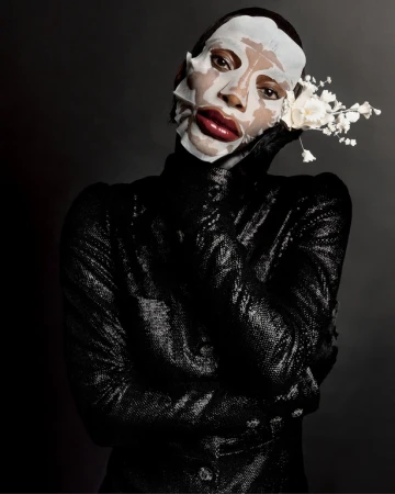 Person in black sequined garment and facial mask poses with white flowers against dark background in beauty editorial photography.