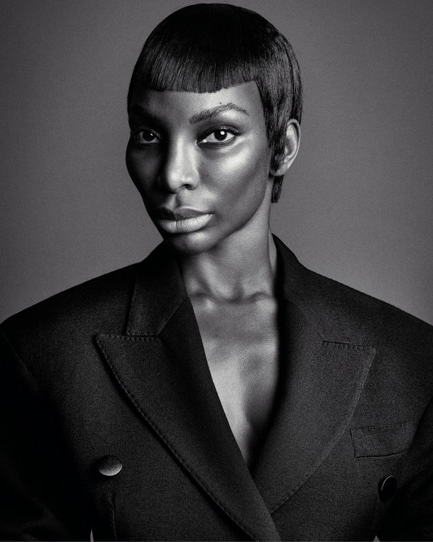 Monochrome portrait of a Black woman with short hair and a black blazer against a plain background in a studio setting.