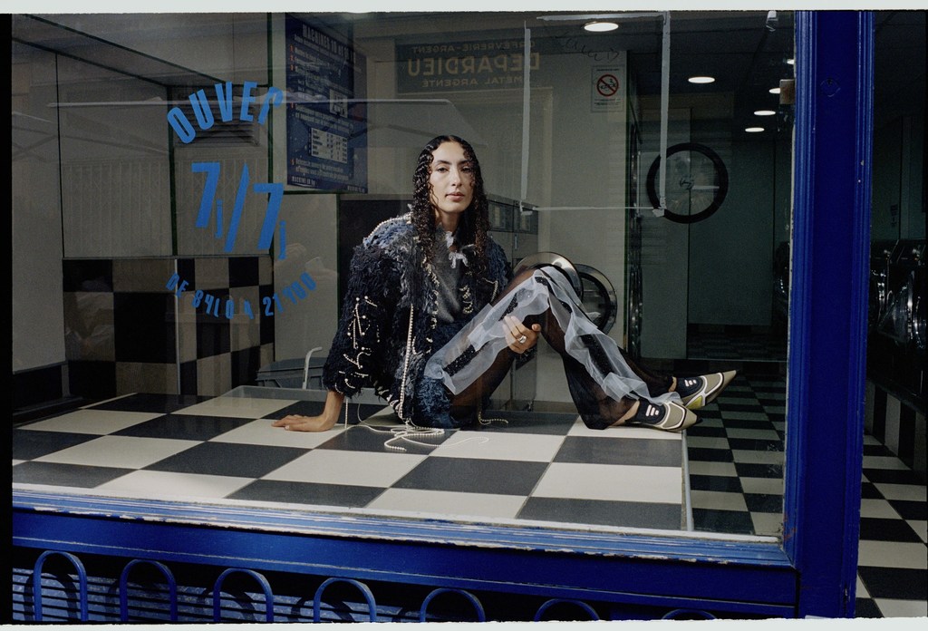 Woman poses in a black, embellished jacket and sheer pants inside a laundromat with blue and white checkered flooring and blue trim.
