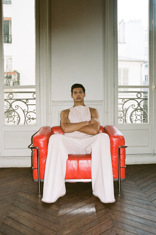 Indoor fashion portrait of a young adult male sitting in a bright red chair, wearing a light pink sleeveless top and matching pants.