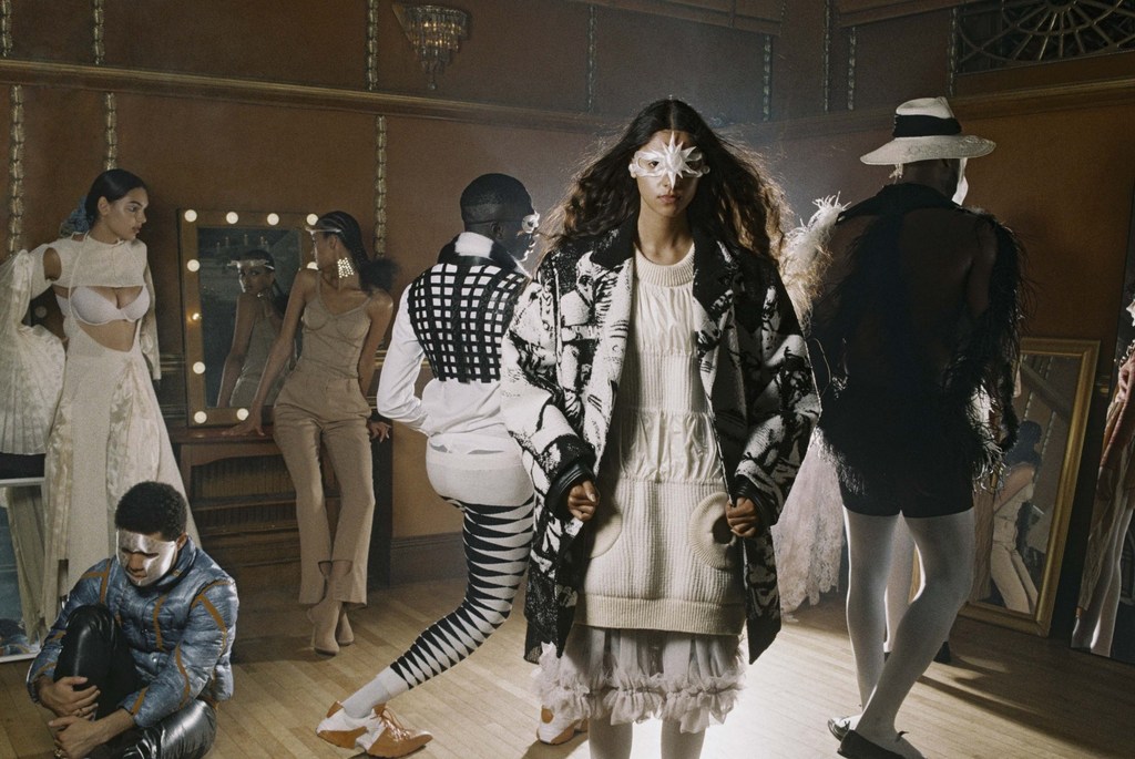 Several fashion models pose in a room with soft lighting, wearing avant-garde clothing and white face makeup in a fashion editorial.