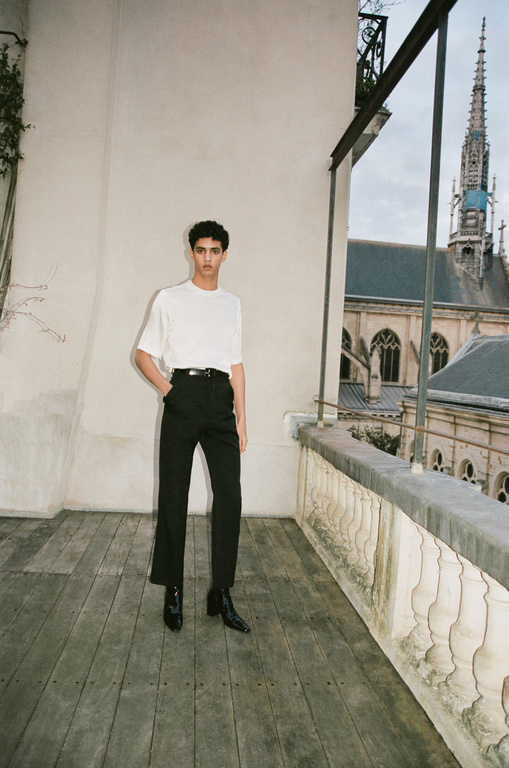 Fashionable adult poses in white tee and black trousers on wood balcony, facing a cathedral against a cloudy sky in outdoor setting.