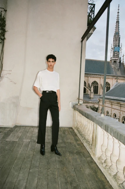 Fashionable adult poses in white tee and black trousers on wood balcony, facing a cathedral against a cloudy sky in outdoor setting.
