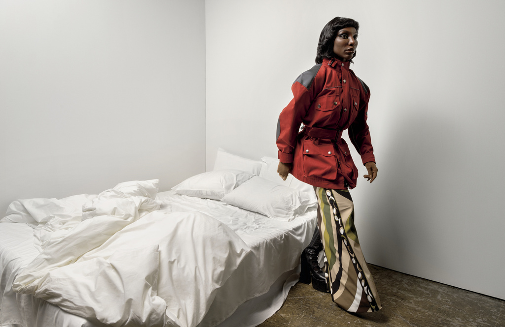 Adult female in a red jacket and patterned pants standing in a stark white interior, bedroom in the background with unmade white bedding.