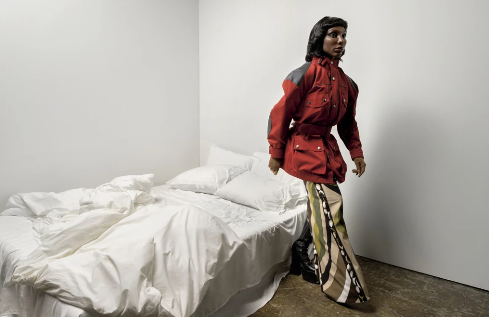 Adult female in a red jacket and patterned pants standing in a stark white interior, bedroom in the background with unmade white bedding.