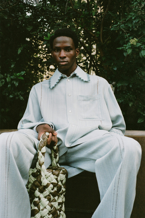 An African American man in a light blue linen suit sits outdoors holding a brown and tan woven bag, with green foliage in the background.
