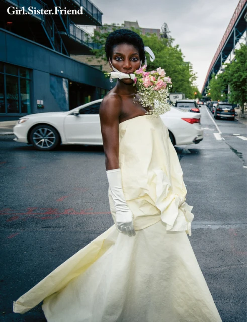 A woman with dark skin and short hair stands on a city street wearing a light-yellow strapless dress, long gloves, and flowers in her mouth.