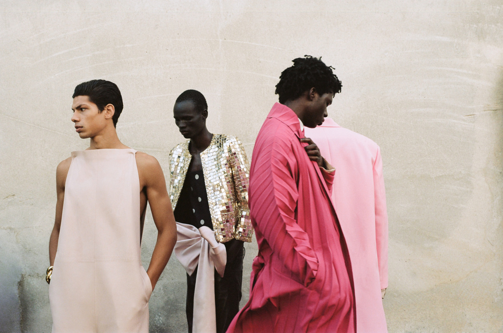 Four dark-skinned male fashion models in pink, gold, and black outfits pose in front of a textured wall in a medium-shot image.