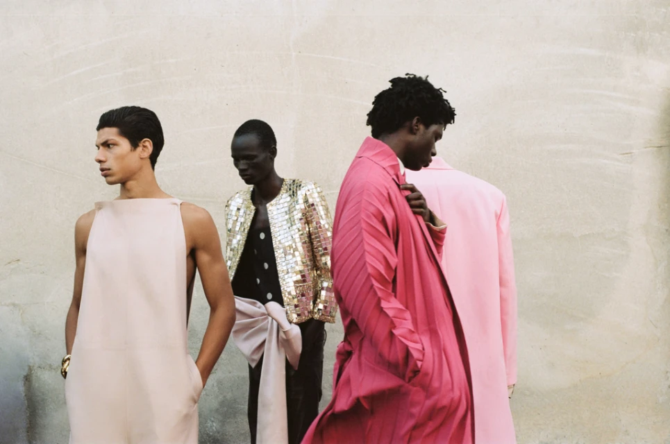 Four dark-skinned male fashion models in pink, gold, and black outfits pose in front of a textured wall in a medium-shot image.