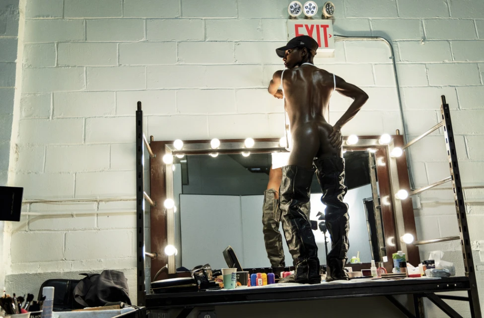 Adult male in hat and high-fashion boots strikes pose backstage in dressing room with lighted mirror and exposed brick wall background.