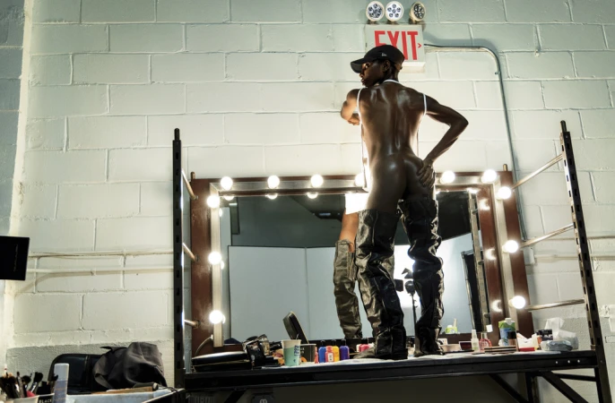 Adult male in hat and high-fashion boots strikes pose backstage in dressing room with lighted mirror and exposed brick wall background.