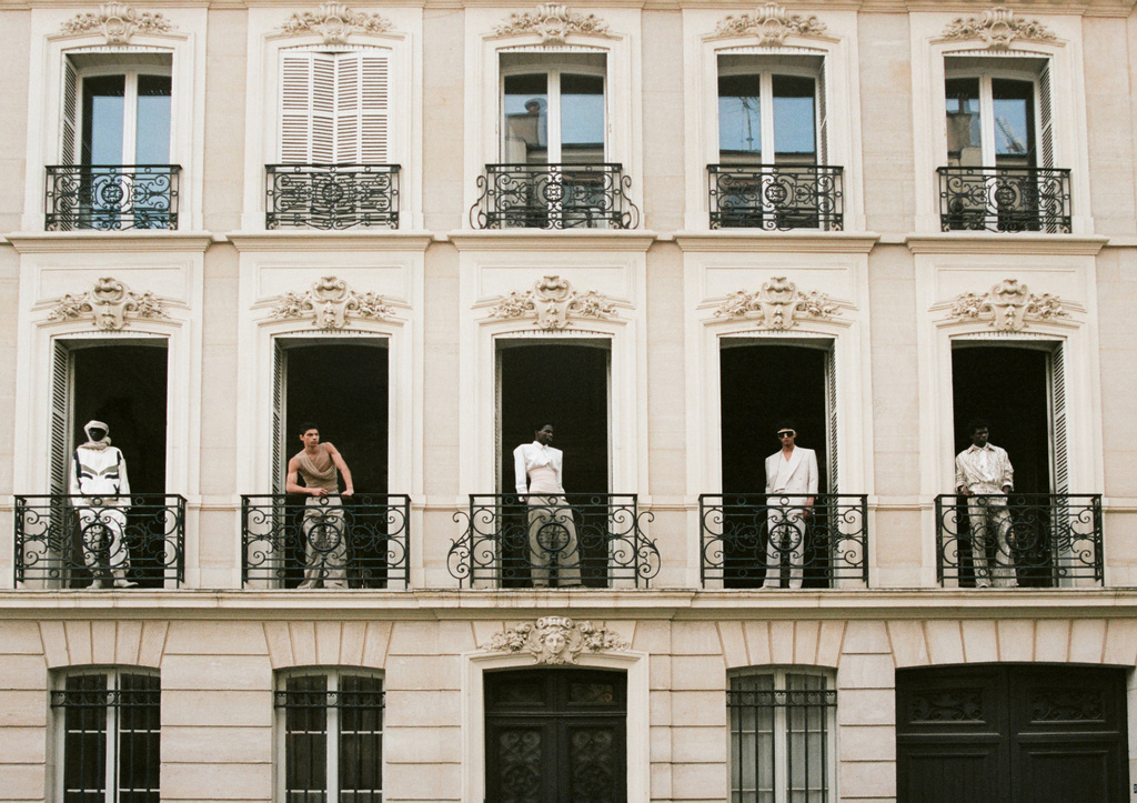 Architectural fashion shot of five models standing in window openings on a building exterior with wrought iron balconies and stone trim.
