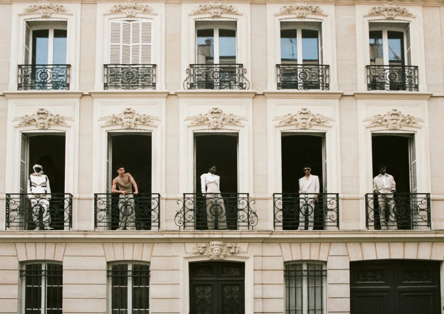 Architectural fashion shot of five models standing in window openings on a building exterior with wrought iron balconies and stone trim.