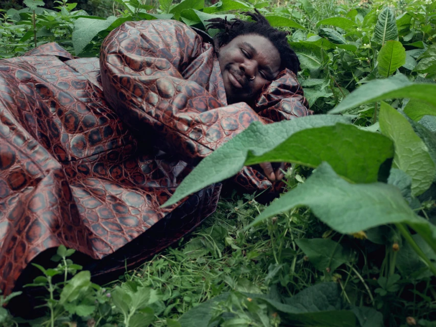 Adult male with dreadlocks lounges in a brown faux-crocodile coat surrounded by green plants, leaves, and grass in an outdoor environment.