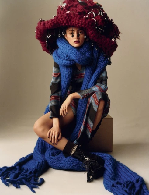 A fair-skinned adult female model poses indoors wearing a large burgundy knit hat, blue knit scarf, plaid dress, and black boots.
