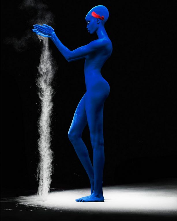 A blue-skinned person stands in profile, pouring white powder from cupped hands in a stark studio shot against a black backdrop.