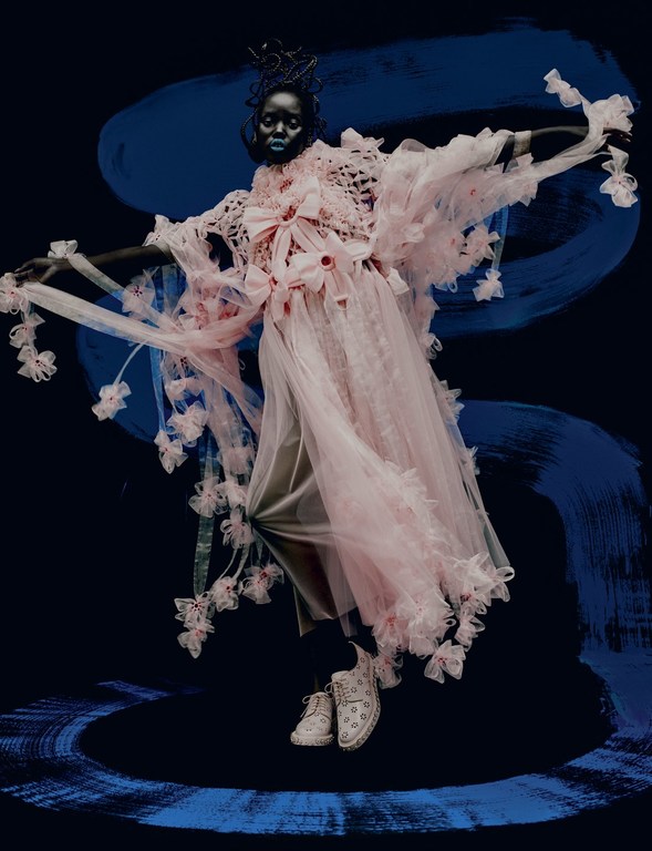 A person with blue skin wearing an elaborate pink tulle gown with white sneakers poses in front of a brushstroke blue background.