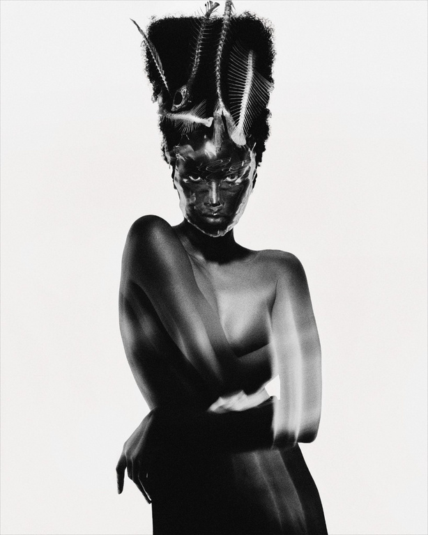 Black woman with sculpted hair adorned with fish skeletons wearing dark makeup against a plain white background