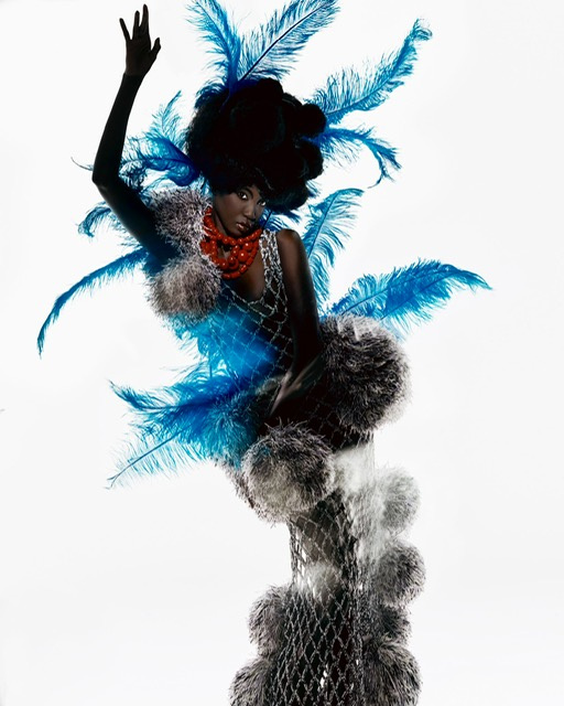 Elegant Black woman poses in ornate fashion costume with blue feathers and a pearl net dress, against a clean white backdrop.