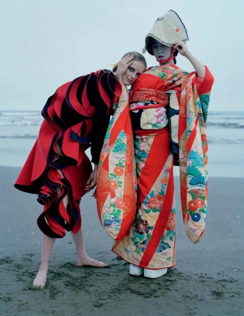 Two women on a beach. One wears a red kimono and straw hat; the other a black-and-red dress. Ocean and sky in the background.