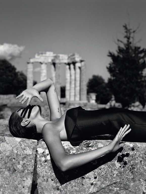 Monochromatic shot of a woman reclining on a stone structure in a black outfit, with a partial building visible in the background.