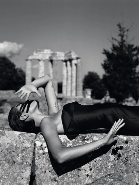 Monochromatic shot of a woman reclining on a stone structure in a black outfit, with a partial building visible in the background.
