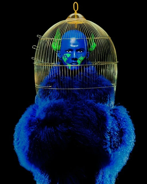 Conceptual image of a person with blue paint in a gold birdcage, wearing a bright blue furry outfit on a black background in studio lighting.