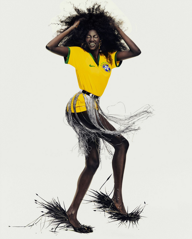 Woman in yellow jersey and fringed skirt poses with joy against a white backdrop in studio lighting. Dark curly hair.