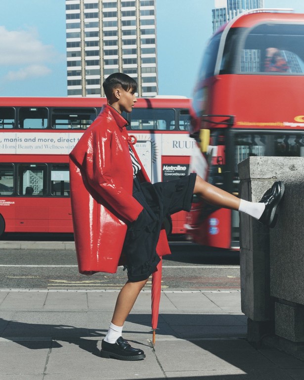 Fashionable woman in red coat poses in city with double-decker bus. Dark hair, black shorts, and umbrella visible in an urban setting.